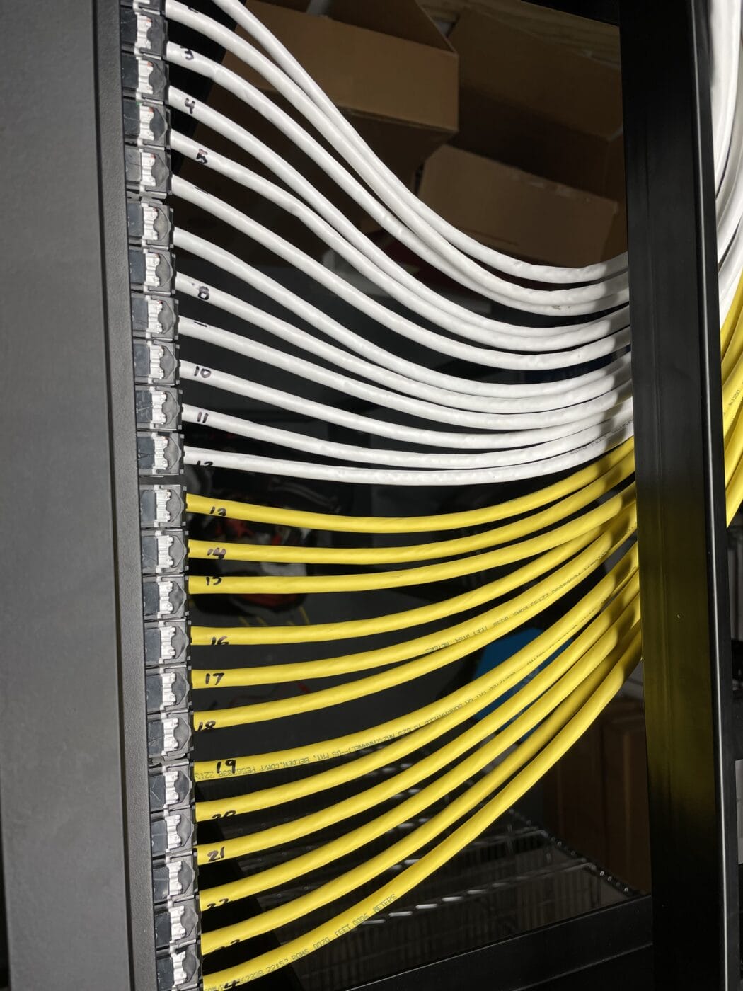 A network patch panel with numbered ports features white and yellow Ethernet cables neatly connected and organized, showcasing professional structured cabling. Cardboard boxes are visible in the background.